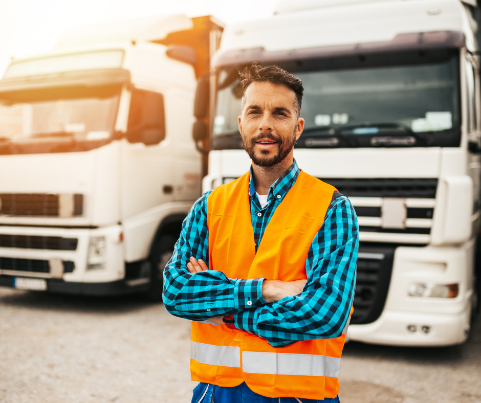 The image shows a confident truck driver standing in front of two white semi-trucks. The driver is wearing a vibrant orange safety vest over a blue plaid shirt, with arms crossed, reflecting professionalism and readiness.