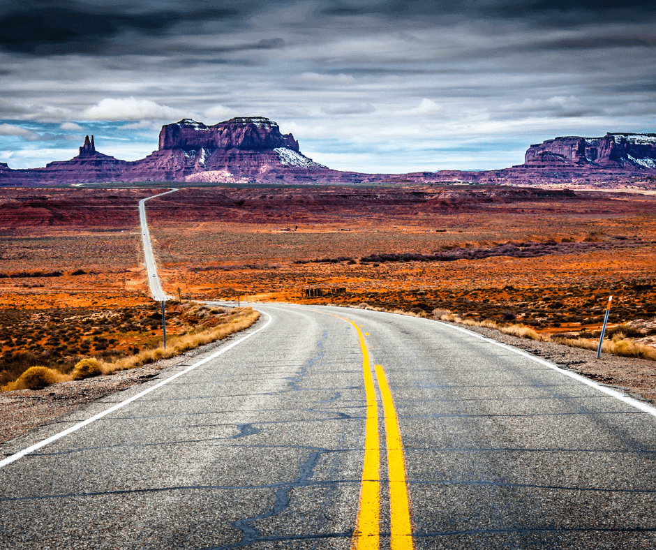 The image shows a long road stretching toward red rock formations under a cloudy sky, likely depicting the American Southwest. The scene conveys a sense of vastness and adventure, with the road symbolizing a journey through open landscapes.