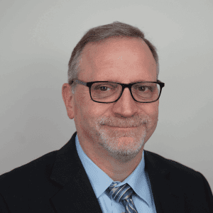 This image depicts Mike wearing glasses, a dark suit, and a light blue dress shirt with a striped tie. He has short, graying hair, a trimmed beard, and a subtle smile while looking directly at the camera. The background is plain, allowing full focus on the individual.