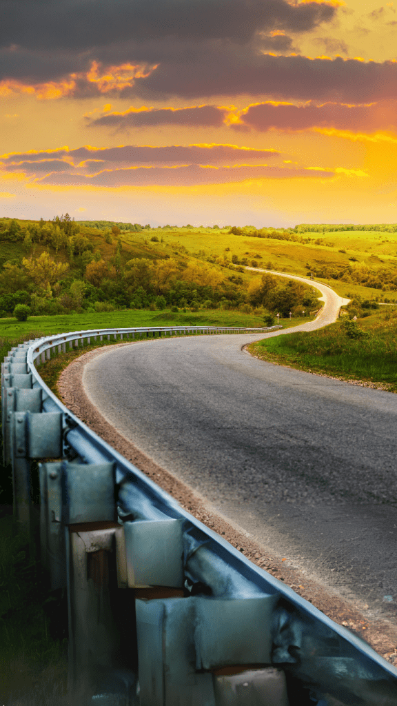 The image shows a winding road with a guardrail, leading through a lush green landscape under a vibrant, colorful sky at sunset. The scene has a serene and picturesque quality, with the road curving into the distance.