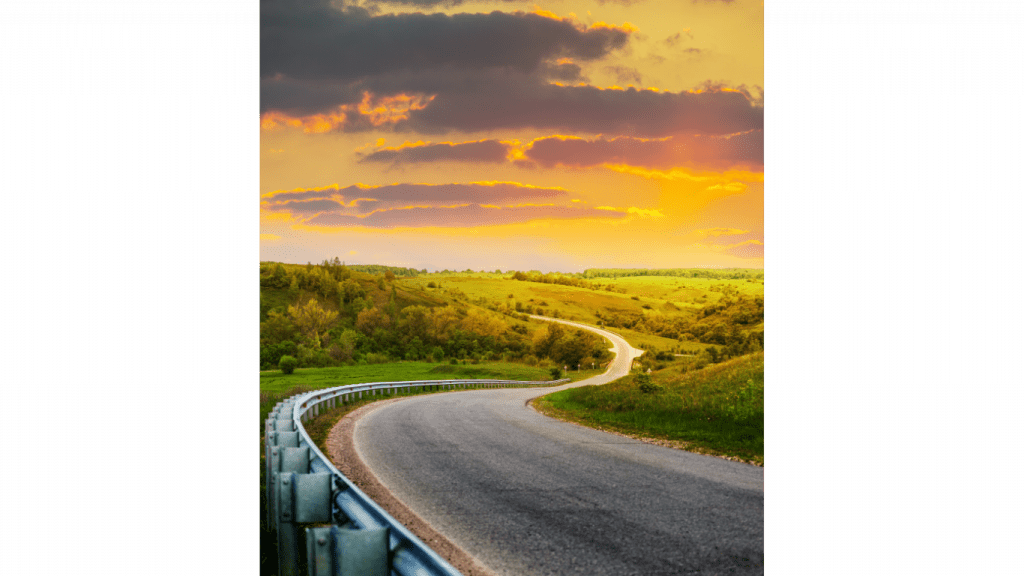 The image shows a winding road with a guardrail, leading through a lush green landscape under a vibrant, colorful sky at sunset. The scene has a serene and picturesque quality, with the road curving into the distance.