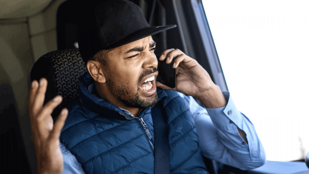 The image shows a frustrated man in a vehicle, talking on a phone with an angry expression. He’s wearing a black cap, a blue vest, and a seatbelt, gesturing with his left hand as he speaks.