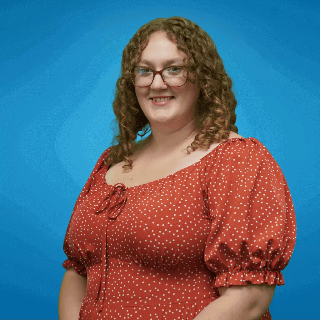 The image shows Taylor with long, curly hair, wearing a poke-a-dot dress. She is smiling at the camera, and her background is a solid light blue.