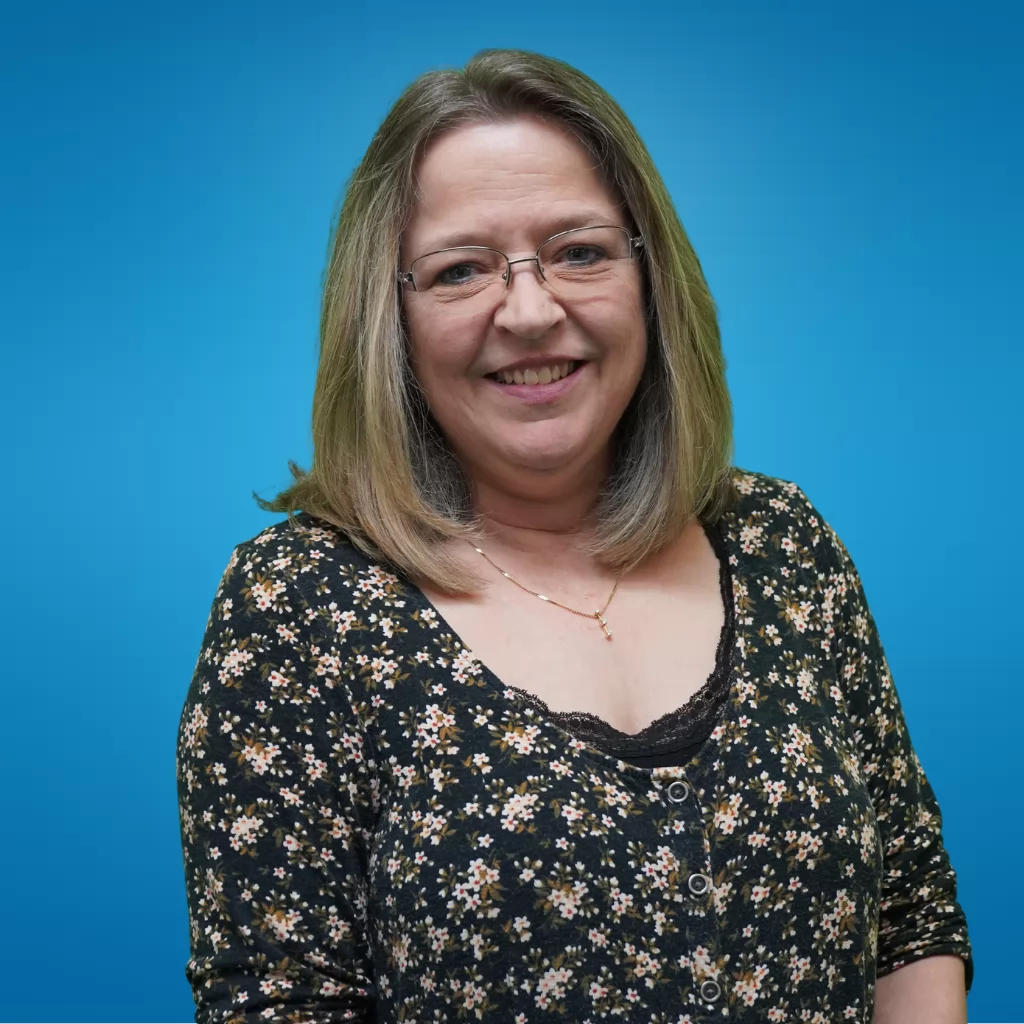 The image shows Erika with short, light brown hair, wearing glasses and a black floral print shirt. She is smiling warmly at the camera, set against a solid blue background.